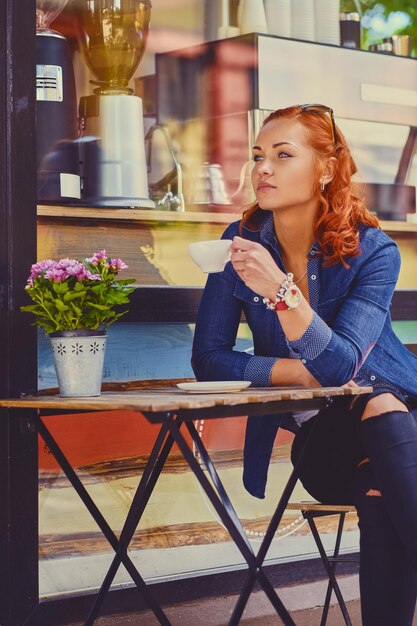 Portrait of redhead female in sunglasses, drinks coffee in a cafe on a street.