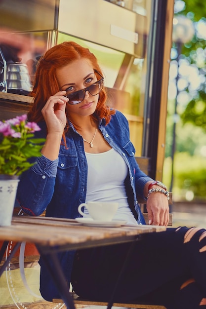 Portrait of redhead female in sunglasses, drinks coffee in a cafe on a street.