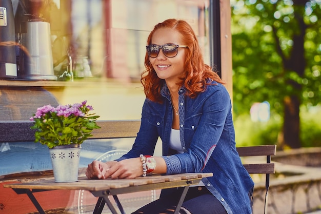Portrait of redhead female in sunglasses, drinks coffee in a cafe on a street.