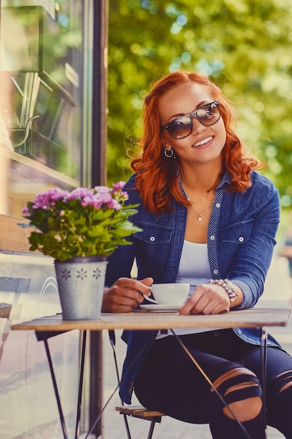 Portrait of redhead female in sunglasses, drinks coffee in a cafe on a street.