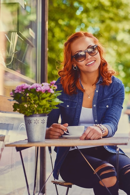 선글라스를 쓴 빨간 머리 여성의 초상화는 거리의 카페에서 커피를 마신다.