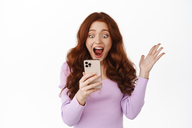 Portrait of redhead girl feeling excited about big promotion on smartphone, reading fascinating news online on mobile phone, scream happy and excited at notification on cellphone