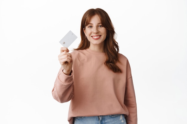 Portrait of redhead girl smiling, showing credit card, advertising bank, special offers or discounts, going on shopping, standing on white.