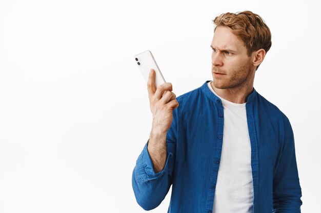 Portrait of redhead guy looks confused at smartphone, hear something strange in mobile phone during call, standing puzzled against white background
