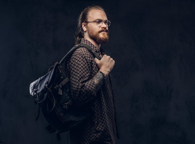 Portrait of a redhead hipster student in glasses dressed in a brown shirt, holds a backpack, posing at a studio. Isolated on a dark background.