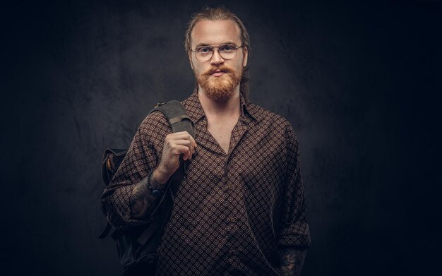 Portrait of a redhead hipster student in glasses dressed in a brown shirt, holds a backpack, posing at a studio. Isolated on a dark background.