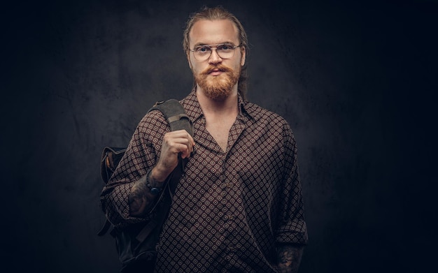 Portrait of a redhead hipster student in glasses dressed in a brown shirt, holds a backpack, posing at a studio. Isolated on a dark background.