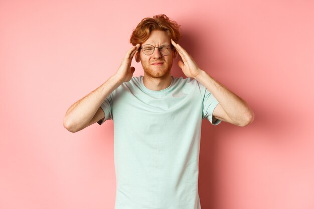 Portrait of redhead man in crooked glasses touching head and feeling dizzy or nauseous having hangov...