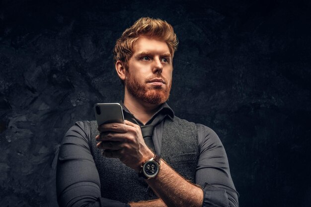 Portrait of a redhead man in formal wear holding a smartphone in studio against a dark textured wall