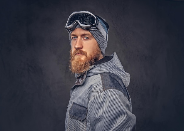 Portrait of a redhead snowboarder with a full beard in a winter hat and protective glasses dressed in a snowboarding coat posing at a studio, looking away. Isolated on a dark textured background.