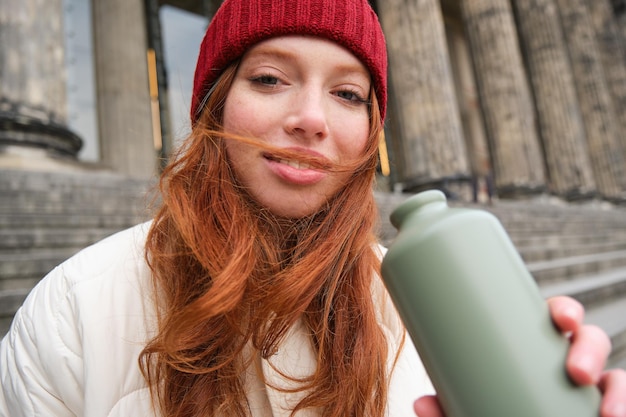 Free photo portrait of redhead woman drinking from thermos sitting on street stairs and enjoys hot drink from f