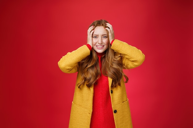 Portrait of redhead woman in panic holding hands pressed to head clenching teeth and frowning puzzled and troubled, standing anxious and concerned freaking out over red background.