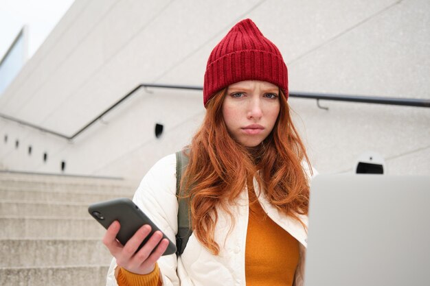 Free photo portrait of redhead woman sits on stairs uses laptop and holds smartphone looks confused and upset a