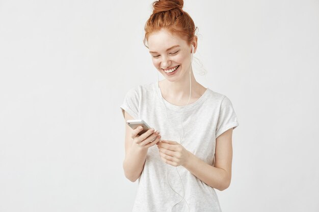 Portrait of redhead woman smiling messaging and listening to streaming music in wired headphones on white wall.