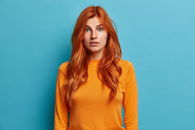 Portrait of redhead young woman looks with wonder and astonishment dressed in casual orange jumper has surpirsed face.