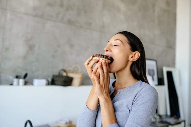 喜んでいる女性の肖像画は、家でおいしいドーナツを食べます。不健康な食品の概念。