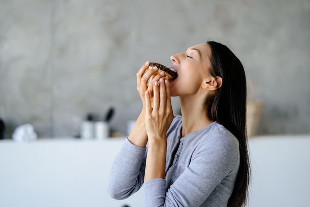 기뻐하는 여성의 초상화는 집에서 맛있는 도넛을 먹습니다. 건강에 해로운 음식 개념입니다.