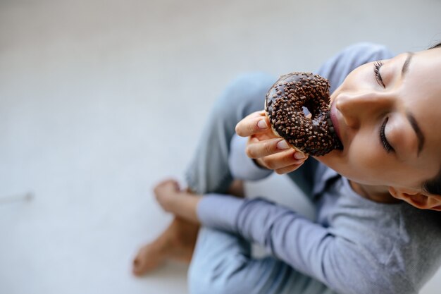 Portrait of rejoicing woman eats tasty donut at home.