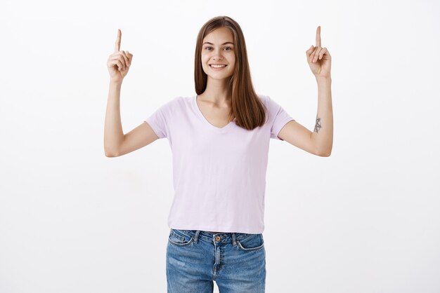 Portrait of relaxed hopeful and deligted cute european woman with tattoo raising index fingers and pointing up smiling with satisfied look