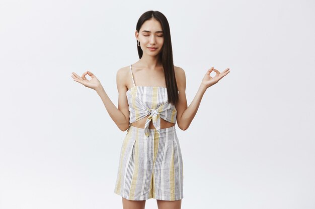 Portrait of relaxed and pleased calm asian woman in summer matching outfit, holding hands spread in zen gesture, smiling, closing eyes while meditating or doing yoga
