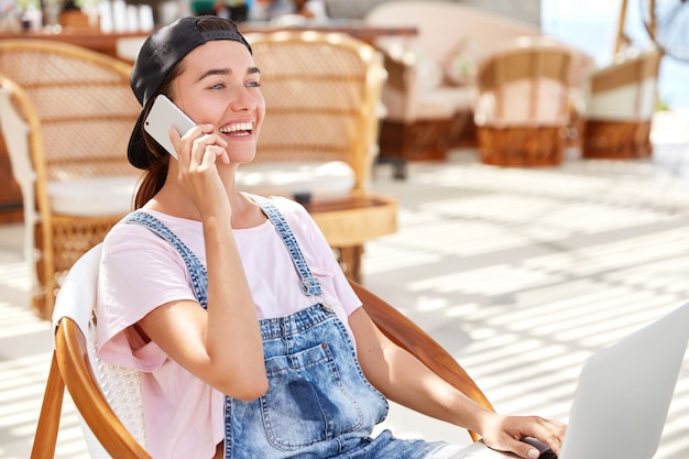 Portrait of relaxed stylish hipster woman in cap and denim overalls, enjoys communication on smart phone while recreats in coffee shop