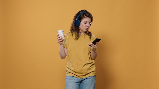 Portrait of relaxing woman holding recyclable coffee cup while using smartphone listening to music on wireless headphones in studio. Person holding mobile phone enjoying online streaming app.