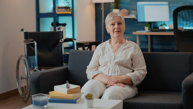 Portrait of retired woman with disability sitting on sofa, next to wheelchair in living room to help with mobility and transportation. Adult enjoying retirement and physical rehabilitation.