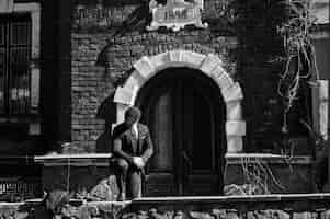 Foto gratuita ritratto di uomo d'affari arabo inglese retrò degli anni '20 che indossa un abito scuro cravatta e berretto piatto in piedi contro la vecchia casa di mattoni 1898 anno