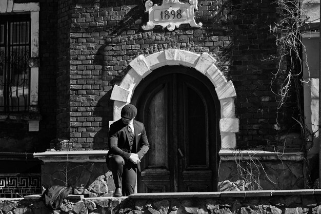 Portrait of retro 1920s english arabian business man wearing dark suit tie and flat cap standing against old brick house 1898 year