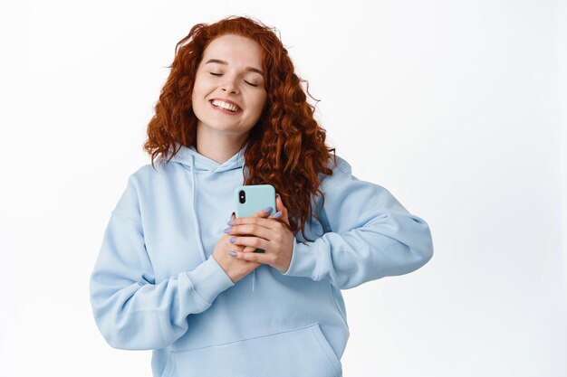 Portrait of romantic smiling girl with ginger curly hair, press smartphone to heart, hugging phone and close eyes delighted, receive good news on cellphone on white