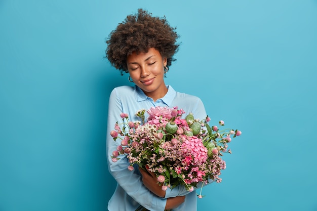 Portrait of romantic young woman embraces pretty flowers, gets bouquet from secret admirer, feels touched, stands with eyes closed, wears blue clothing