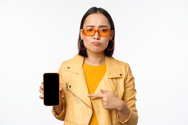 Portrait of sad asian woman in sunglasses pointing finger at mobile phone app interface showing smartphone application standing over white background