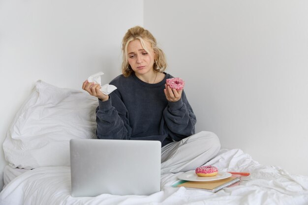 Portrait of sad crying young woman staying at home sitting in bed with doughnut and comfort food