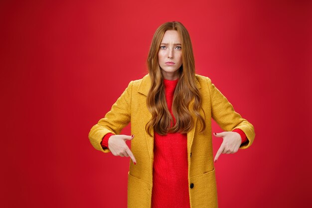 Portrait of sad and displeased moody girlfriend asking opinion with concerned upset expression pointing down looking focused and serious with unhappy face at camera over red background.