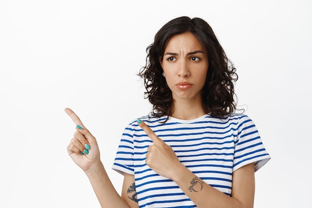 Portrait of sad frowning girl with curly hair, pierced nose, pointing and looking left with disappointed expression, sulking upset, complaining, standing  on white