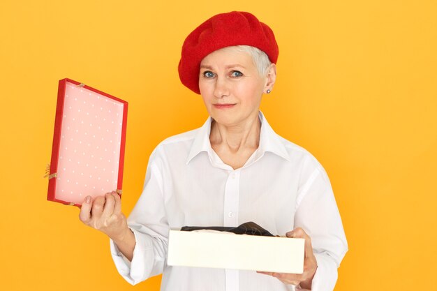 portrait of sad frustrated mature retired female in red beret holding box, unpacking present on Valentine's Day