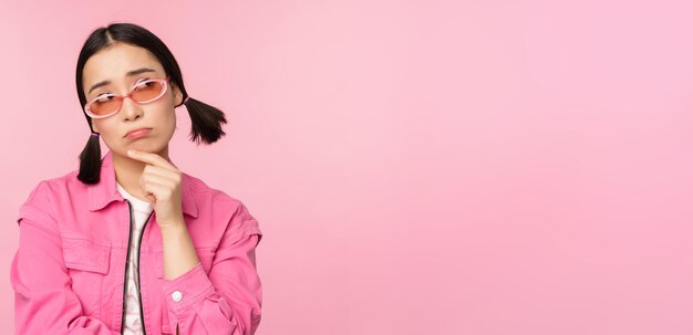 Портрет грустной и мрачной азиатской девушки, дующейся от разочарования, стоящей расстроенной на фоне розовой студии
