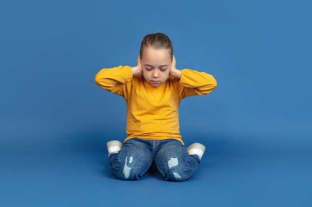 Portrait of sad little girl sitting isolated on blue studio background. How it feels to be autist. Modern problems, new vision of social issues. Concept of autism, childhood, healthcare, medicine.