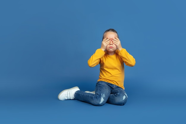 Portrait of sad little girl sitting isolated on blue studio background. How it feels to be autist. Modern problems, new vision of social issues. Concept of autism, childhood, healthcare, medicine.