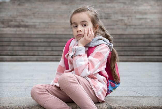 Портрет грустной маленькой девочки с рюкзаком, сидящей на лестнице. Обратно в школу.