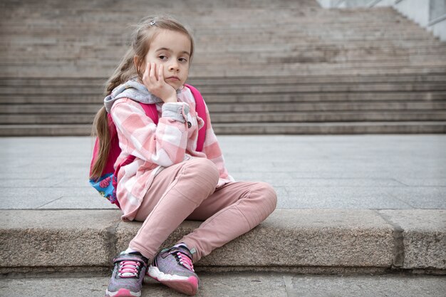 Портрет грустной маленькой девочки с портфелем на спине, сидящей на лестнице. Обратно в школу.