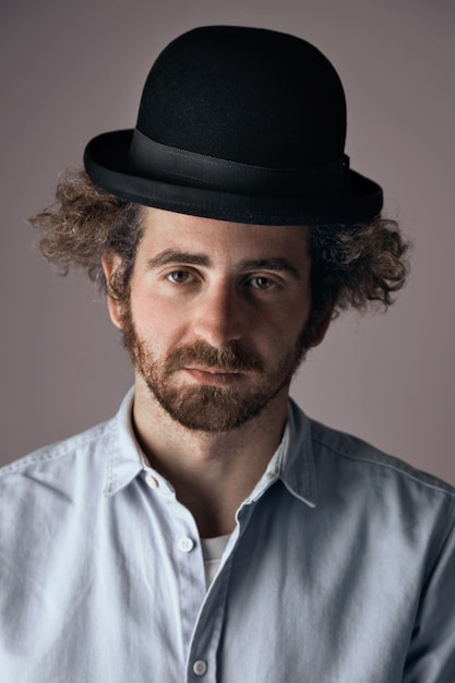 Portrait of a sad looking young bearded jewish man with curly hair wearing a funny black bowler hat and light denim button up t-shirt isolated on light gray.