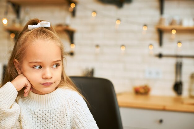 Portrait of sad moody seven year old female child in cozy warm sweater spending cold winter day at home alone, keeping hand under her chin, having thoughtful upset facial expression, looking away