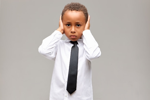 Portrait of sad unhappy African American boy in school uniform having upset depressed facial expression, covering ears with hands, can't stand parents fighting. Body language, reaction and feelings