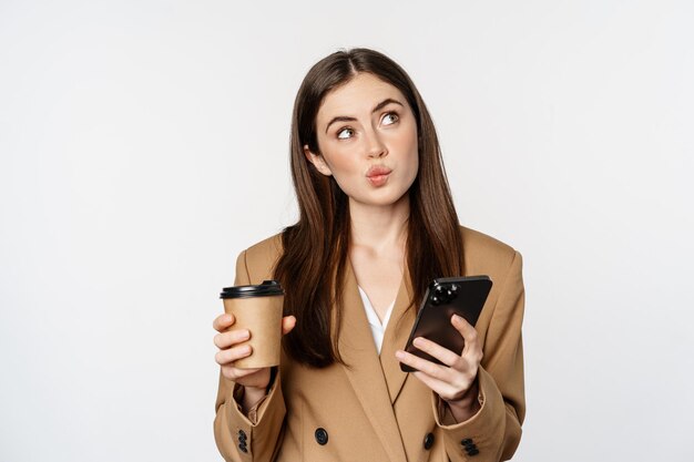 Портрет продавщицы корпоративной женщины, пьющей кофе на вынос и читающей смартфон, смотрящей на мо...