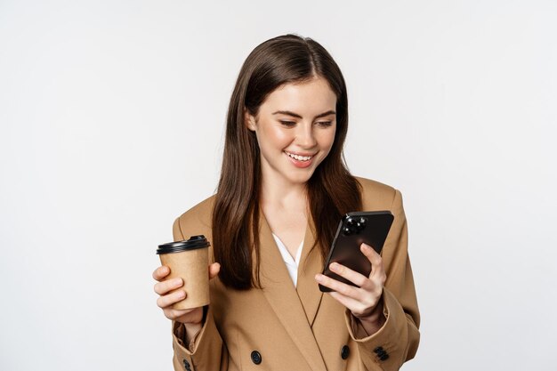 Портрет продавщицы корпоративной женщины, пьющей кофе на вынос и читающей смартфон, смотрящей на мо...