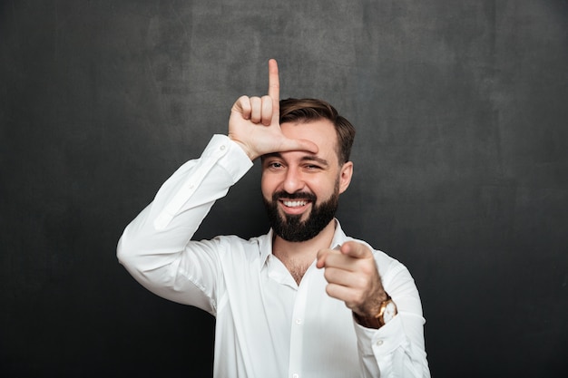 Free photo portrait of sarcastic man showing loser sign on his forehead and pointing on camera with smile, mocking or humiliate over graphite wall