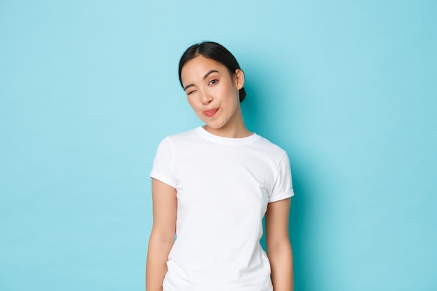 Portrait of sassy and attractive asian girl in white casual t-shirt, showing tongue and winking coquettish while standing over blue wall.