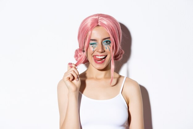Портрет нахальной привлекательной девушки в розовом парике, одетой в одежду для Хэллоуина, улыбаясь и подмигивая в камеру, стоя.