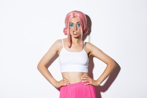 Free photo portrait of sassy attractive woman with pink anime wig, bright makeup, showing teeth and looking confident, celebrating halloween, standing over white background